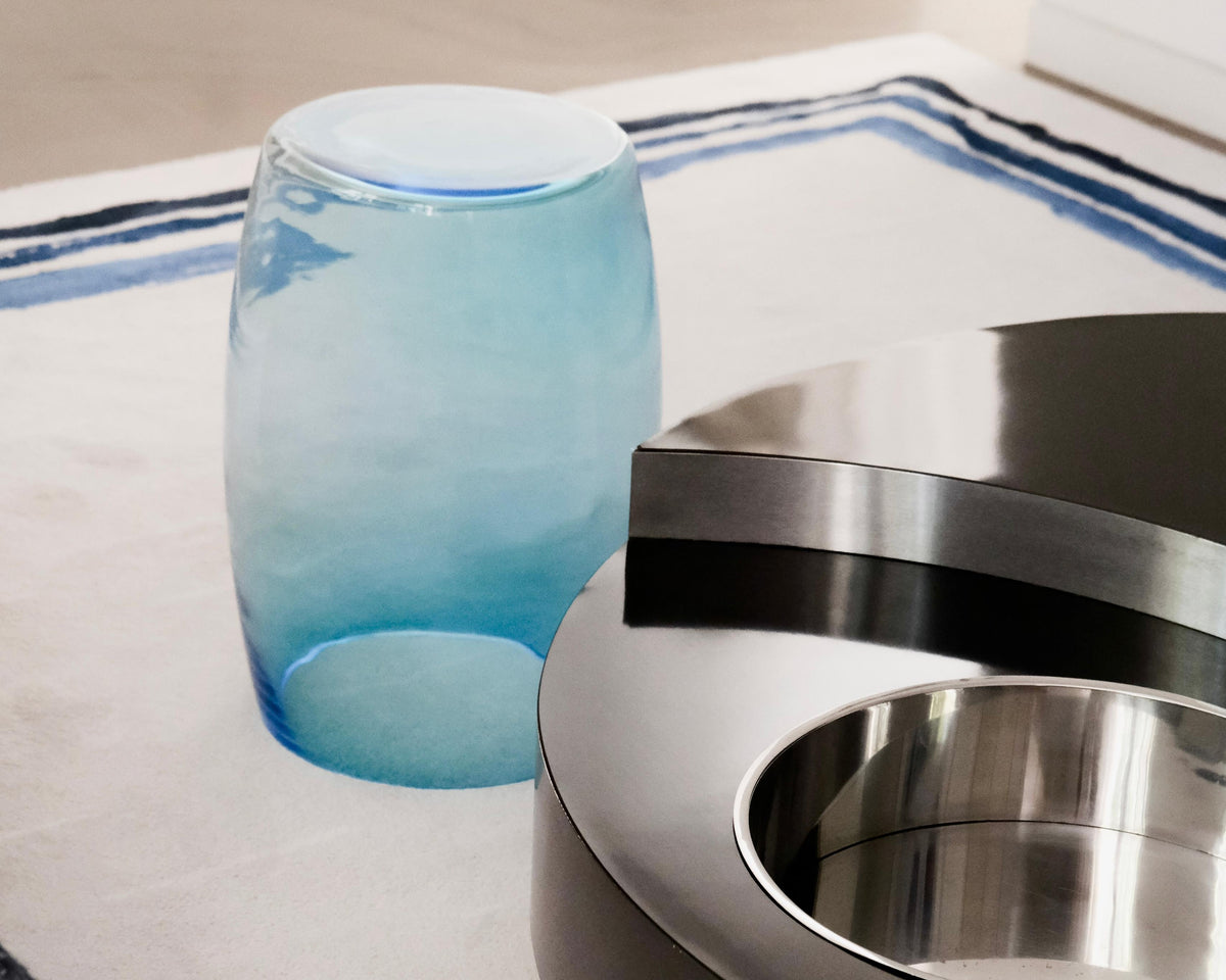 Tavolino Acqua Mare 50 beside a stainless-steel side table, showing aquamarine transparency and light refractions in a contemporary interior.